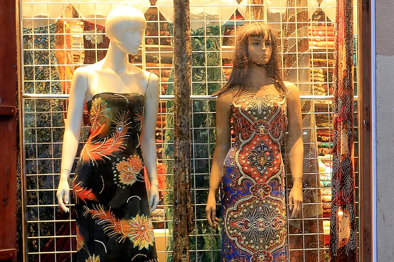 A shop window in the Bur Dubai cloth souq, two mannequins in printed and embroidered dresses standing in front of bolts of cloth behind a wire grid.