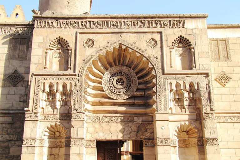 The carved stone front of the al Aqmar mosque, with a fluted hood like a shell over the doorway, a round pierced medallion at its centre, carved niches and rosettes on either side and an inscription band along the top.