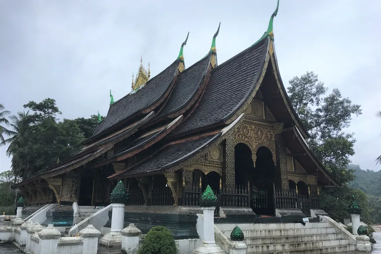 The hall of Wat Xieng Thong in Luang Prabang, its dark tiled roofs layered low toward the ground, with green finials, a gilded spire on the ridge and a gold carved porch above white steps and balustrades.