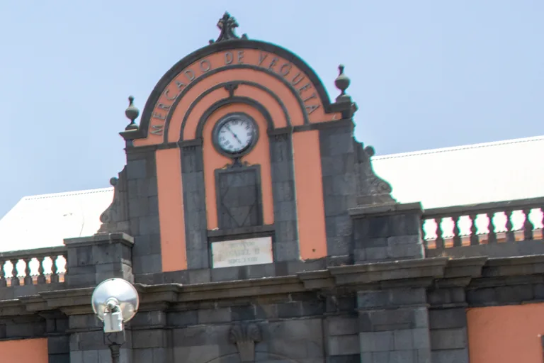 The upper front of the Vegueta Market, a curved gable of salmon plaster and dark stone lettered Mercado de Vegueta round a clock, with a stone balustrade, a street lamp and a pale roof behind, against a clear sky.