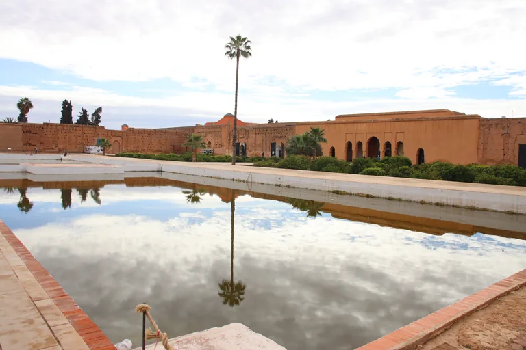 The long rectangular pool of the El Badi Palace reflecting the clouds and a tall palm tree, with the high earthen walls and arched ochre pavilions of the ruined palace beyond.