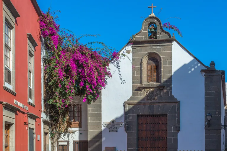 The white hermitage of San Antonio Abad, a grey stone bell gable with a bell and a cross above a tall wooden door, with purple bougainvillea spilling over the wall at the left beside a red house front.