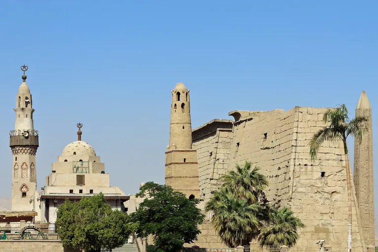 The Abu Haggag mosque high on Luxor Temple, with its white minaret and dome on the left and its older mud brick minaret against the temple pylon, palm trees and the tip of an obelisk on the right.