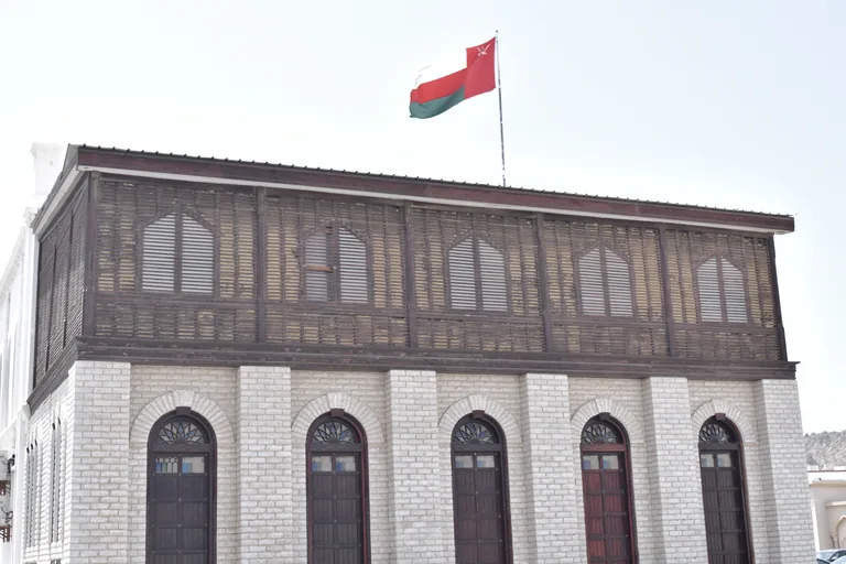 The front of Bait al Baranda, a two storey house whose upper floor is wrapped in a screened wooden balcony, above white brick walls and tall arched wooden doors, with the Omani flag flying on the roof.
