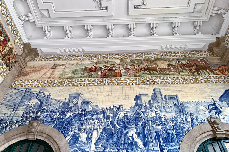 Blue and white tile panels of a royal procession in the hall of São Bento Station, beneath a coloured frieze.