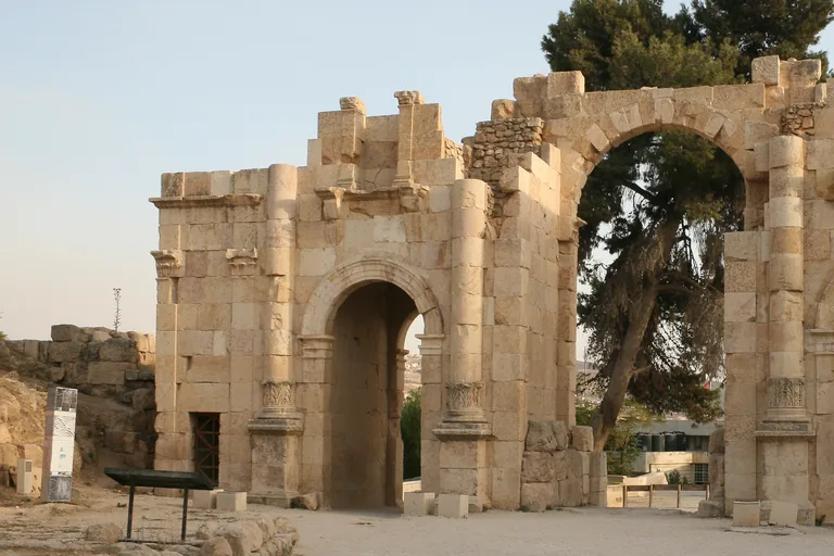 The South Gate of Jerash, a ruined gateway of pale squared stone with a tall arched passage, a large central arch framing a pine tree and carved half columns on its front, on a gravel forecourt in evening light.