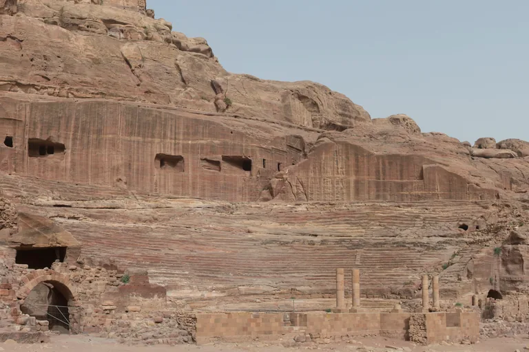 The Petra theatre, curved rows of seats cut into the mountainside below a cliff of opened tomb chambers, with the rebuilt stage front and a few standing columns in the foreground.