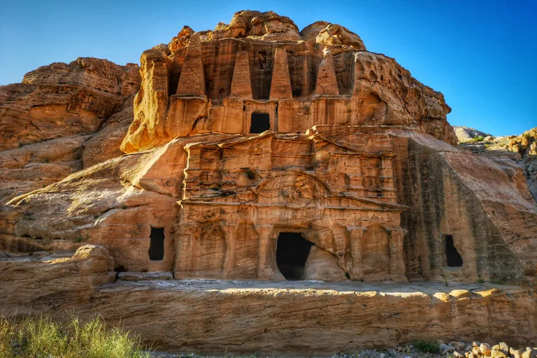 The Obelisk Tomb at Bab as Siq, four tall obelisks and a niche cut into the top of a sandstone outcrop, above the columned and gabled front of the triclinium with its dark doorway, in low morning sun under a blue sky.