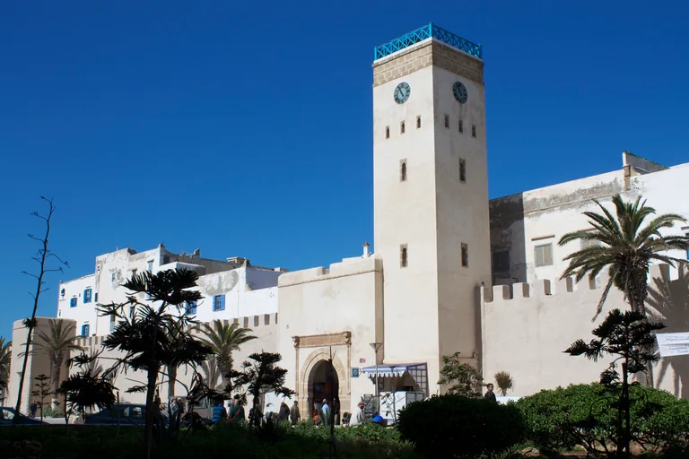 The white clock tower over the arched gate in the crenellated Kasbah wall, with palms and garden shrubs in front under a clear blue sky.