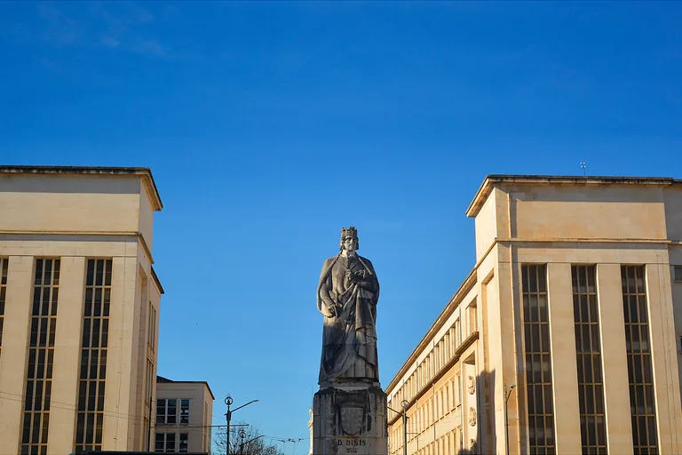 The statue of King Dinis on a tall pedestal between two pale stone faculty buildings under a clear sky.