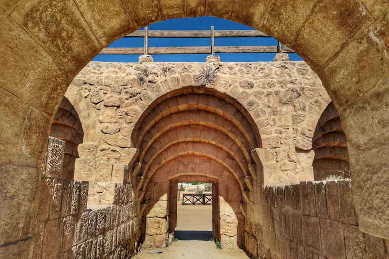 Looking through a stone archway at the hippodrome's starting gates, a tunnel of stepped arches between rough stone walls leading to a doorway onto the sandy arena, with a wooden rail above the wall.
