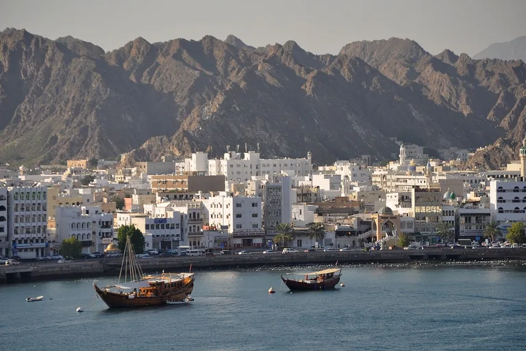 Two wooden dhows moored in the bay of Mutrah in front of the corniche wall and the white town, with domes and minarets among the buildings and bare jagged mountains behind.