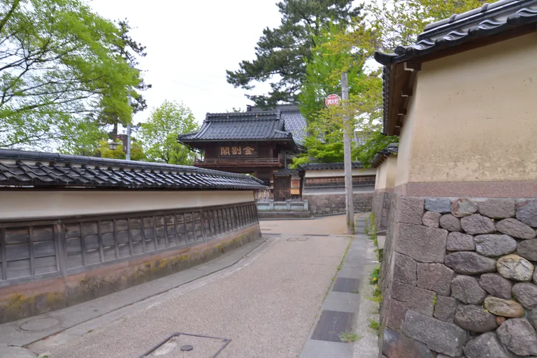 A narrow gravel lane between long plastered temple walls, tiled along the top and set on stone, in Kanazawa, bending toward a wooden gate of two storeys with a balcony rail and a name board, under a tall pine and fresh green trees.