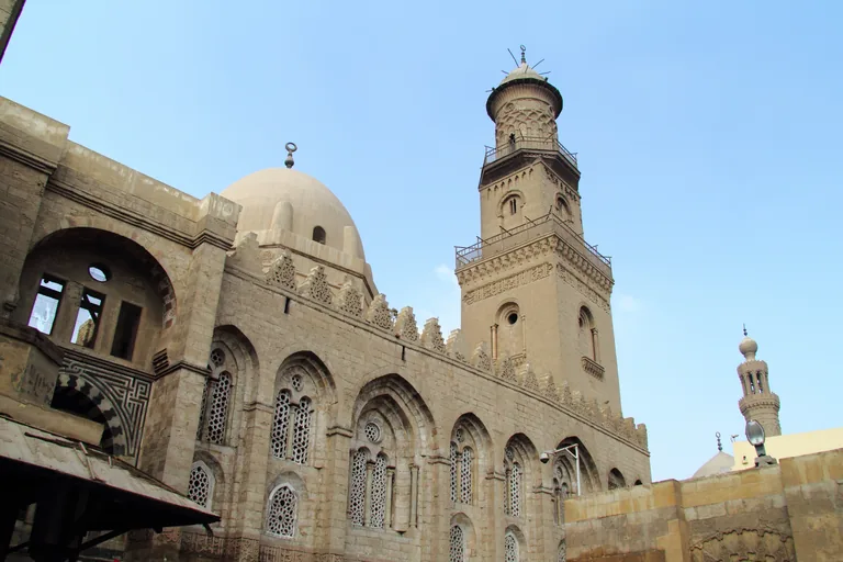 The street front of the Qalawun complex on al Muizz Street, a long stone wall of pointed arched window bays with a dome behind and a tall carved minaret, under a pale blue sky.