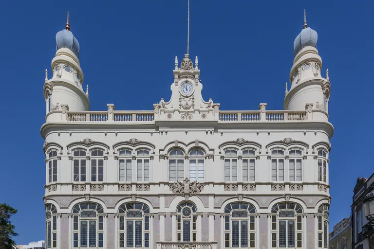 The upper front of the Gabinete Literario, white and cream with tall arched windows, carved balustrades and a clock in the central crest, between two towers crowned with grey onion shaped domes under a deep blue sky.