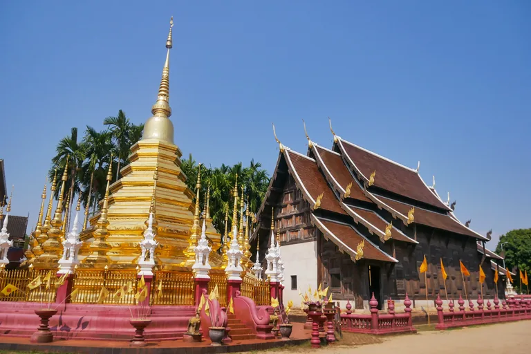 The dark teak hall of Wat Phan Tao in Chiang Mai, its brown tiled roofs stepping down in tiers with golden finials, beside a gilded chedi ringed by smaller spires and palms, behind a low red balustrade on sandy ground.