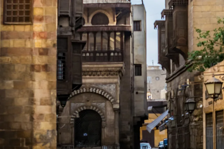 The Sabil Kuttab of Abd al Rahman Katkhuda at the fork of al Muizz Street, its grilled water window under a striped arch with a wooden loggia above, between old stone house fronts with wooden window screens.