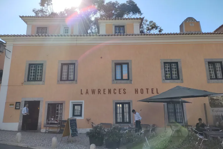 The ochre front of Lawrence's Hotel with its name in large letters, framed windows and a street terrace with tables and umbrellas.