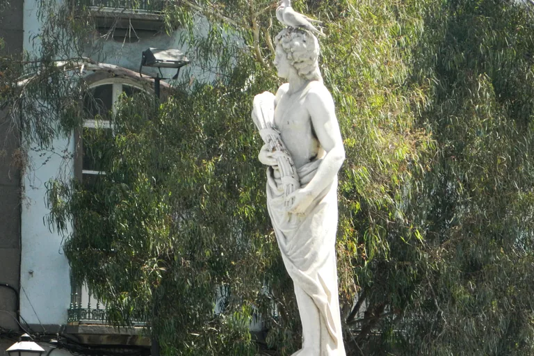 A white marble statue of a draped figure holding a sheaf of wheat, the figure of Summer, with two doves on its head, against the hanging leaves of a tree and a pale house front.