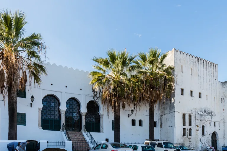 The white crenellated front of Dar el Makhzen on the Kasbah square, with arched windows, a stairway, tall palms and cars parked along the wall.