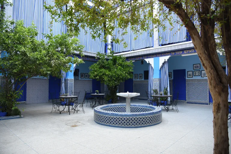 The courtyard of the Lazama synagogue, with a blue and white tiled fountain, café tables, trees, blue doors and a Hebrew inscription running along the blue and white walls.