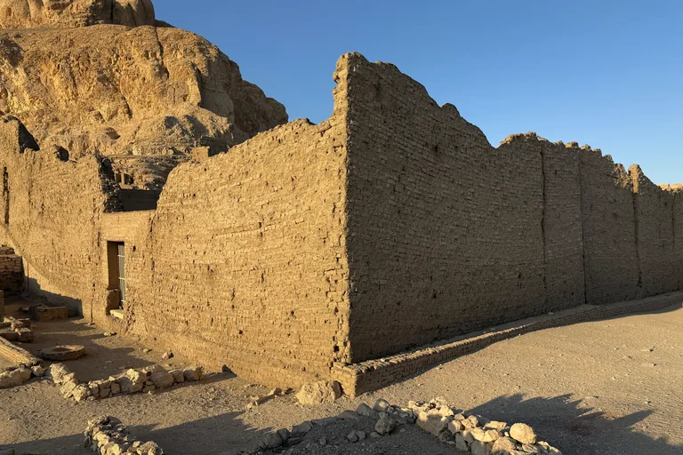 The tall mudbrick enclosure wall of the Hathor temple at Deir el Medina in low evening sun, its corner and long side above a packed earth path, with the cliffs of the Theban mountain rising behind.