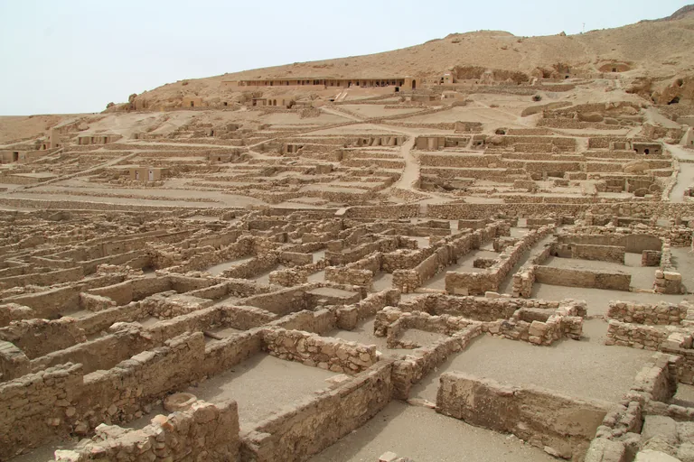 The excavated village of Deir el Medina, rows of low stone house walls and lanes filling the valley floor, with terraced tomb chapels and small pyramids on the slope behind and a long arcaded excavation house near the top.