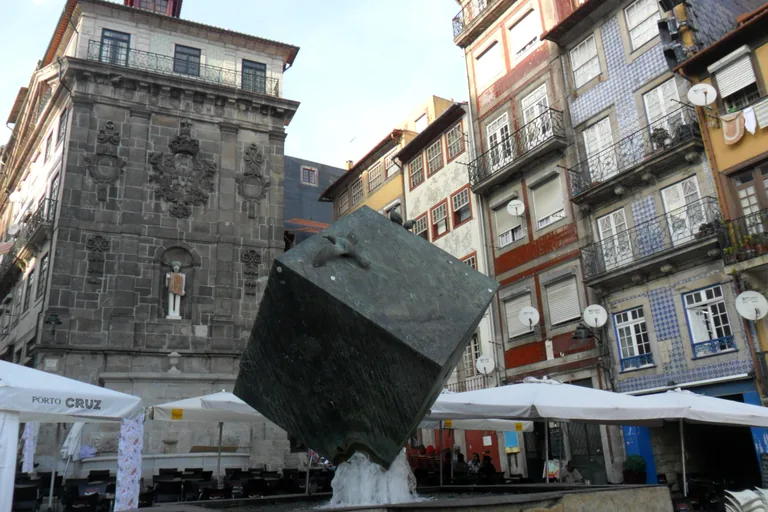 The dark metal cube fountain in Ribeira Square in front of tall tiled houses and café umbrellas.