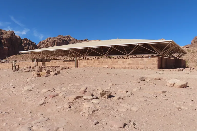 The low sandstone walls of the Petra Church under its wide modern shelter roof on a stony slope, with rocky hills behind under a clear blue sky.