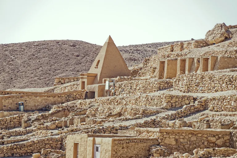 A restored mudbrick pyramid chapel with a small niche on the terraced hillside of the workmen's cemetery, rock cut chapel doorways in a row on the right and dry stone terrace walls below.