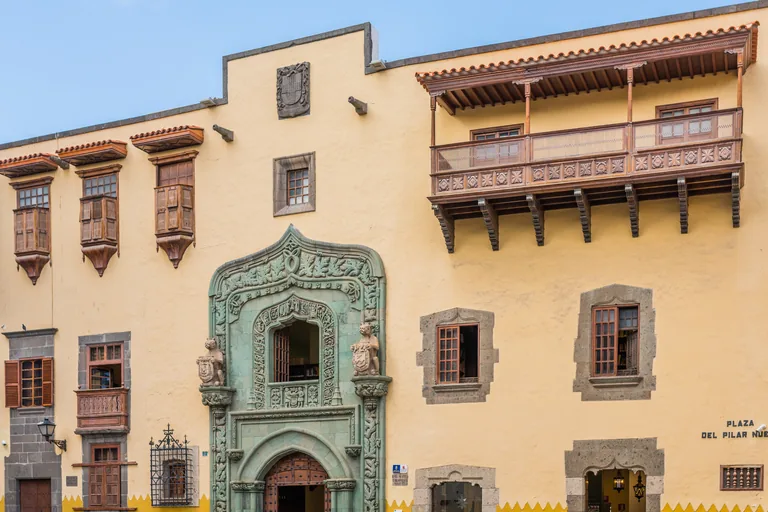 The long yellow front of the Casa de Colón, with carved wooden balconies under tiled eaves, small windows in stone frames and a tall doorway carved in green stone in the middle.