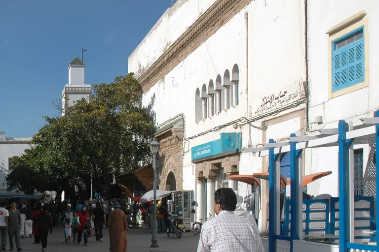 Place Moulay Hassan looking toward the square minaret of the Kasbah Mosque behind a large tree, with people walking past white buildings, café awnings and a blue railing.