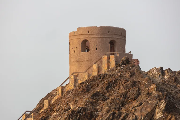 A round watchtower of plastered stone on the rocks of the Riyam headland, reached by a stone stair with a metal rail, with an old cannon in one of its arched openings.