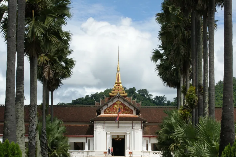 The white front of the Royal Palace in Luang Prabang with its slender golden spire and a carved gilded pediment, framed by tall palm trees, with a wooded hill behind and white cloud in a blue sky.