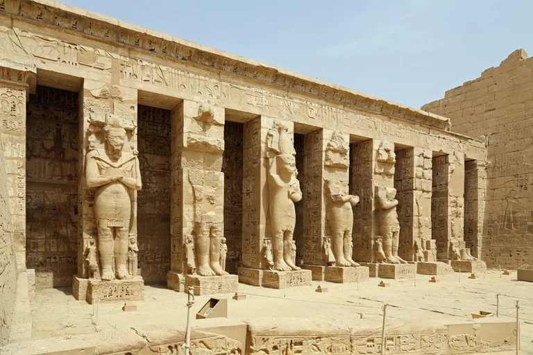 The north side of the court behind the pylon at Medinet Habu, a row of square pillars with giant standing statues of the king, the fuller ones on the left, behind a rope barrier.