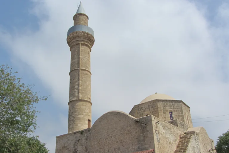 The stone mosque of Agia Sofia in Paphos with its tall round minaret capped by a metal cone, a dome on an eight sided drum and an outside stair climbing the wall, under a cloudy sky.