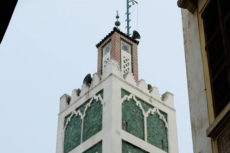 The square minaret of the Grand Mosque of Tangier, faced with green tiles and white carved panels, with merlons and a small lantern on top against a pale sky.