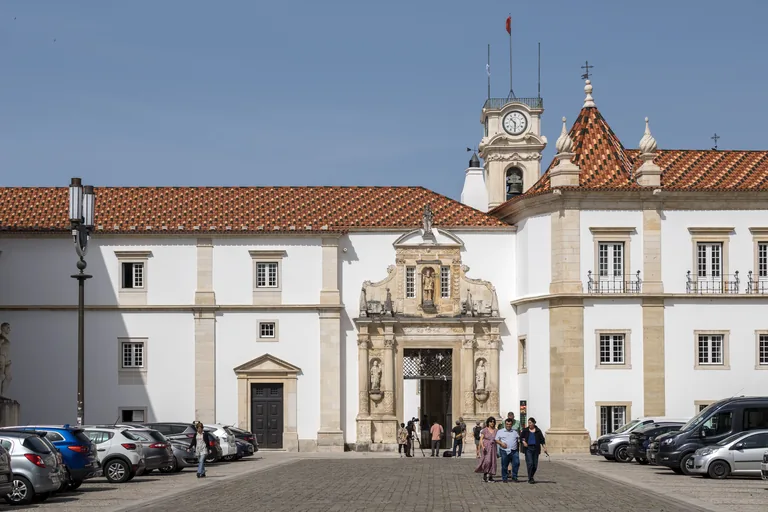 The Porta Férrea gateway in the long white palace wall, with the clock tower rising behind it and cars parked in the square.