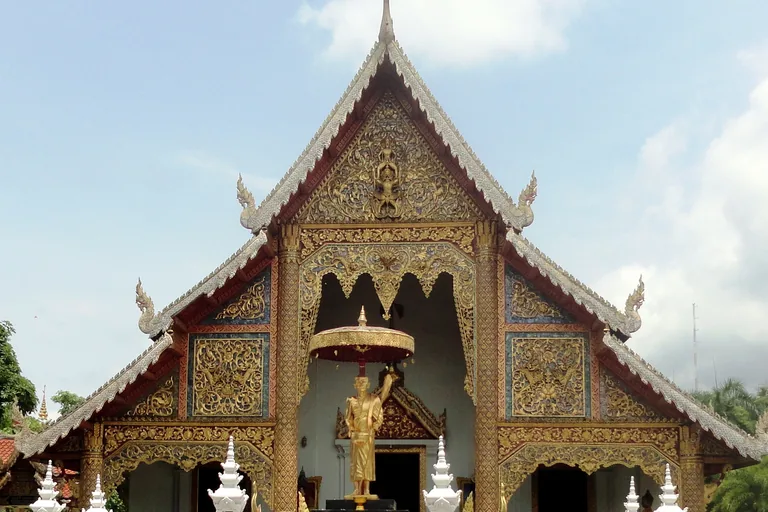 The front gable of the main hall of Wat Phra Singh in Chiang Mai, richly carved and gilded, with silver serpent finials along its tiered roofs and a gilded standing figure under a parasol before the porch.
