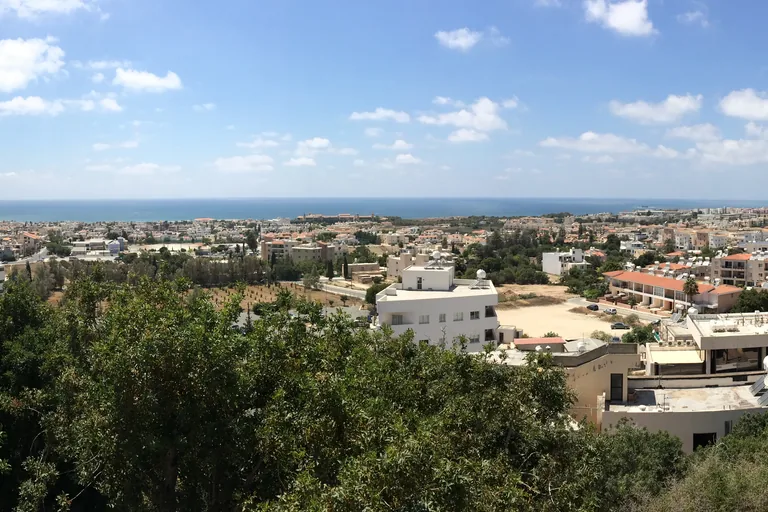 The view from the cliff top of the upper town of Paphos over treetops to the white buildings of the lower town on the plain and the blue sea along the horizon.