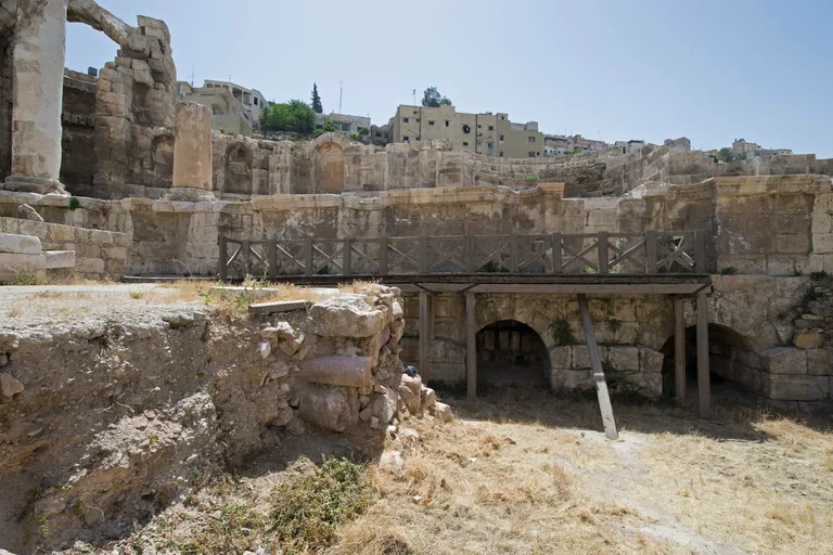 The Nymphaeum in Amman seen from inside its fenced site, ruined walls with curved niches and a standing column, a wooden walkway on posts in front of vaulted openings and dry grass in the foreground, with houses above.