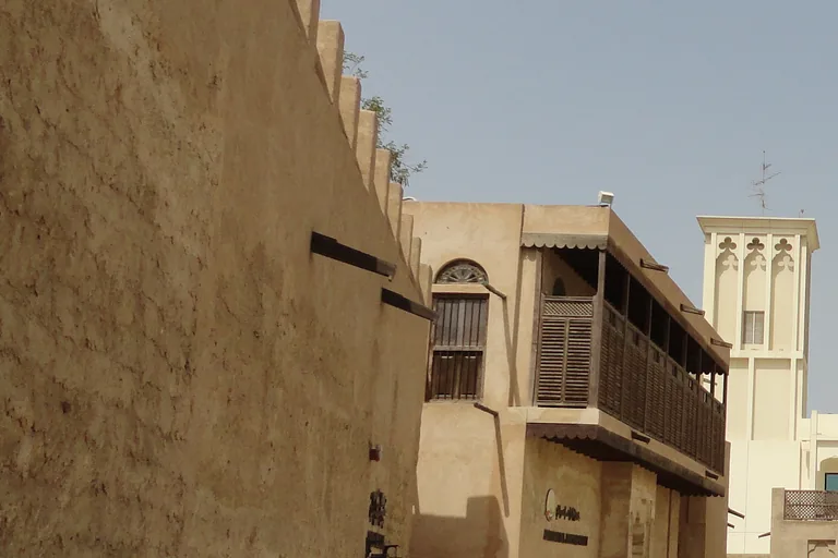 A paved lane in Al Fahidi between a long crenellated wall of plastered stone and a two storey house with a screened wooden balcony, with a pale wind tower rising beyond.