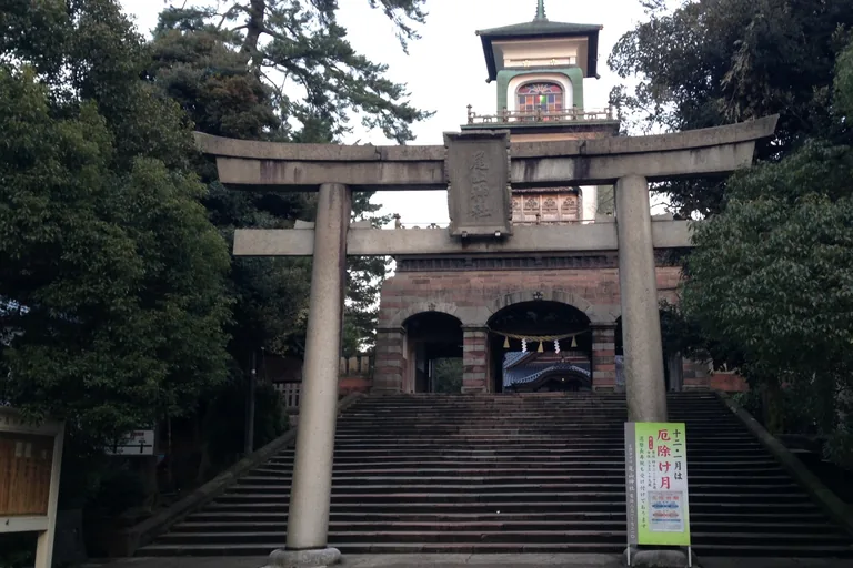 A tall stone torii at the foot of a broad flight of stone steps in Kanazawa, and above the steps a gate of three round arches faced in stone with two smaller white storeys on top, the upper one set with coloured glass, between dark trees.