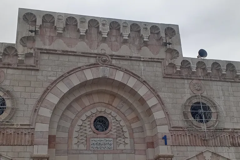 The entrance front of the Grand Husseini Mosque in Amman, a tall arch of alternating pale and pink stone around a carved panel with an Arabic inscription, round windows and shell shaped crenellations along the roofline.