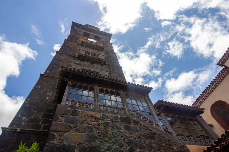 The tower of La Concepción seen from its foot, dark volcanic stone rising to a clock and balconies, above a long glazed wooden gallery under tiled eaves, with clouds in a blue sky.
