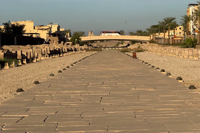 The paved Avenue of Sphinxes running toward a low arched road bridge with a coach crossing it, rows of sphinx bases on both sides and apartment blocks and palm trees beyond the walls.