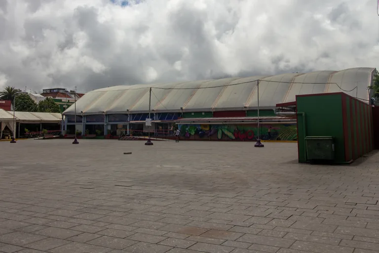 The provisional market hall on the Plaza del Cristo, a long low building under pale curved tent roofs with a painted mural of vegetables along its front, across a wide paved square under heavy cloud.