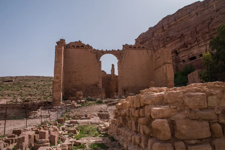 The standing walls of Qasr al Bint with its great arch, seen across fallen blocks and the low wall of the sacred precinct, with the cliff and trees on the right.