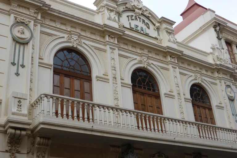 The upper front of the Teatro Leal, carved cream plaster with round medallion portraits, arched wooden windows behind a balustraded balcony, and the theatre's name in green letters under the crest.