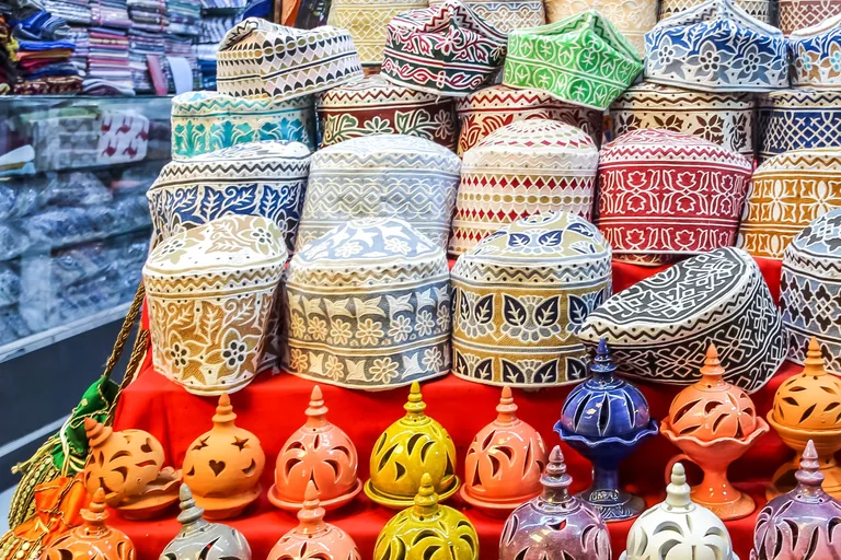 Embroidered Omani caps stacked in rows on a red cloth in a shop of the Mutrah souq, with clay incense burners in many colours lined up in front of them.